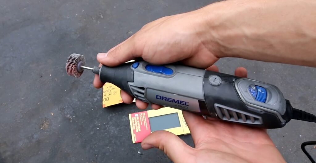 A hand holds a Dremel tool with a wire brush attachment, with sandpaper pieces nearby on a pavement background