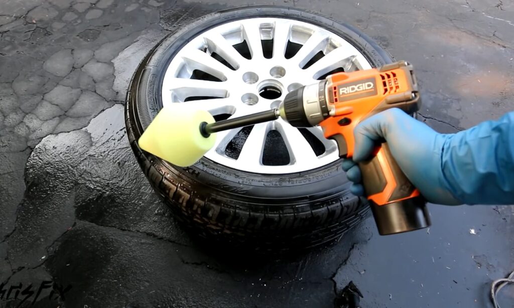 A gloved hand holds an orange RIDGID power drill with a foam attachment, poised over a wet car wheel on cracked pavement
