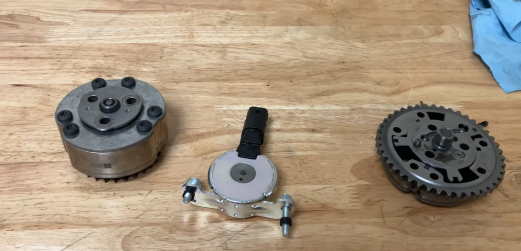Three mechanical components are arranged on a wooden surface: a cylindrical piece, a silver disk with a black plug, and a gear-like structure