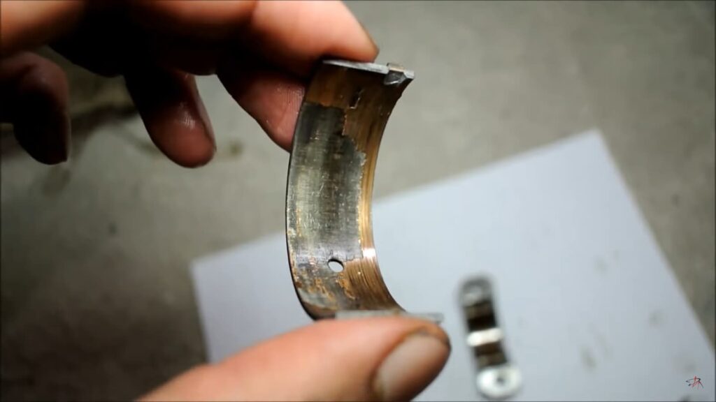 A hand holds a worn-out engine bearing with visible signs of damage, with another part on a white surface in the background.