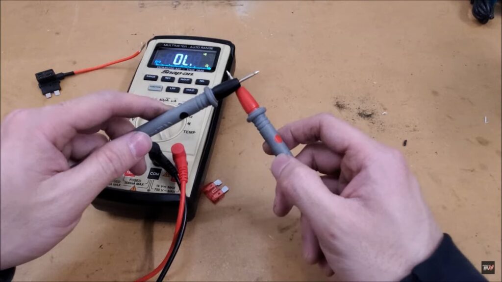 Hands holding multimeter probes near a digital multimeter, with a fuse tap and red fuse on a wooden background